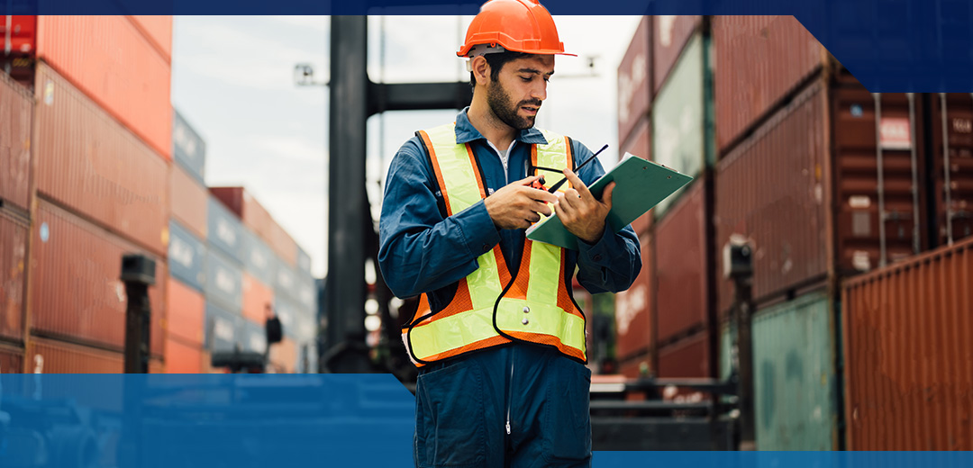 A man in a hard hat and vest in a cargo yard looks at a clipboard