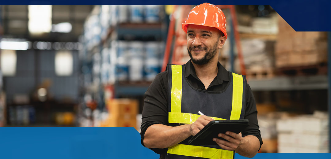 A man in a warehouse wears an orange hard hat and a safety vest while holding an iPad