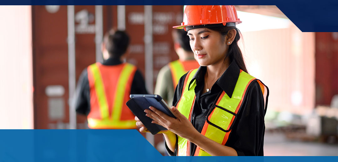 A female wearing an orange hard hat a yellow safety vest looks at a tablet